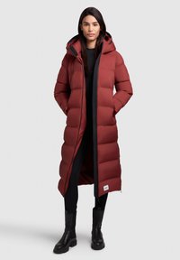 Rød pufferjakke med hætte, der har en knælang design med quiltet tekstur, sorte detaljer og sidelommer. Iført over mørke leggings og støvler.
