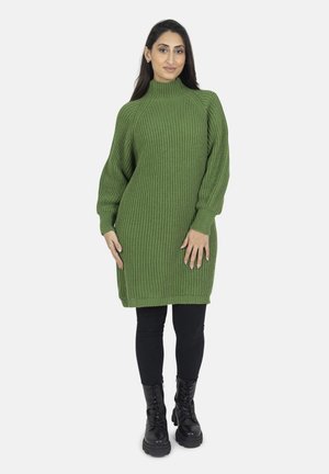 Kvinde iført en grøn ribstrikket sweaterkjole, sorte leggings og sorte snørebånds-kampstøvler, stående mod en hvid baggrund.