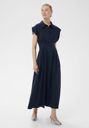 Nő, aki tengerészkék, ujjatlan, midi hosszúságú, galléros és gombos felsőrésszel rendelkező ruhát visel, valamint ezüstszínű, nyitott orrú, magas sarkú papucsokat.