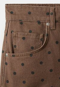 Material din denim maro cu buline negre, detaliind zona buzunarului frontal, bucla pentru curea și nituri metalice.