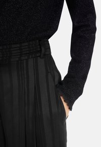 Schwarze plissierte Hose mit vertikalen Streifen, kombiniert mit einem dunklen, funkelnden Langarm-Oberteil mit Rüschenmanschetten.