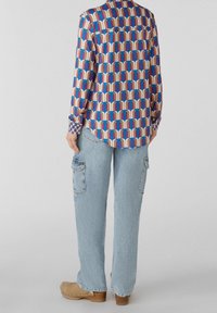 Camicia multicolore a fantasia in tessuto morbido con maniche lunghe e orlo arrotondato, abbinata a jeans larghi di un blu chiaro e zoccoli di legno.