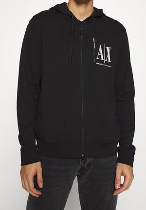 Schwarzer Zip-Hoodie mit Kapuze und Kordeln, mit weißem "A|X Armani Exchange" Logo auf der oberen linken Brust, getragen zu dunklen Jeans.
