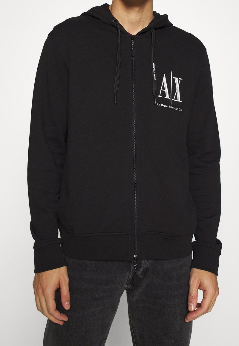 Sweat à capuche noir zippé avec cordons, avec le logo blanc "A|X Armani Exchange" sur la partie haute gauche de la poitrine, porté avec un jean foncé.