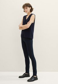 Top sin mangas de canalé en color navy con escote en V, combinado con jeans ajustados de mezclilla oscura y zapatos negros con cordones sobre un fondo neutro.