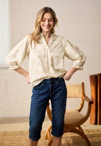 Femme souriante, mains sur les hanches, portant un chemisier crème à motifs floraux et un jean court bleu foncé, debout à l'intérieur près d'une chaise en bois.