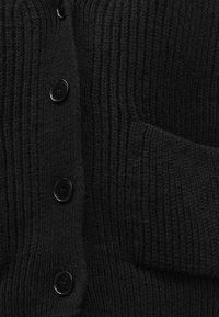 Svart strikket cardigan med vertikale riller, fem store svarte knapper og en fremre lomme. Myk tekstur med et koselig utseende.