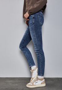 Jeans skinny in denim blu scuro con una fila verticale di borchie decorative sulla cucitura esterna. Indossati con sneakers alte beige e bianche.
