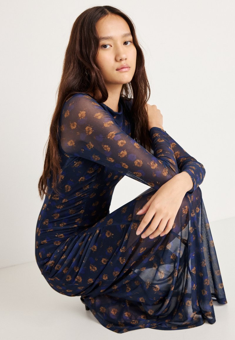 Průsvidné námořnické šaty s dlouhými rukávy, zdobené květinovým vzorem v zlatých tónech. Přiléhavý střih s draped siluetou a jemnou texturou.
