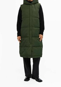 Lange, mit Kapuze versehene Parka in dunklem Grün, gepolstertes Design mit Knopfverschluss und seitlichen Taschen. Über einem schwarzen Pullover und schwarzen weit geschnittenen Hosen getragen.
