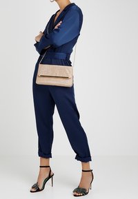 Námornická kombinéza s ohrnutými rukávy, v kombinaci se zlatou třpytivou kabelkou a černými vysokými podpatky se zeleným akcentem a dekorační mašlí.