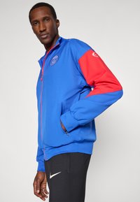 Férfi kék és piros Paris Saint-Germain kabátban, cipzáros zsebekkel és fekete Nike nadrágban, egy sima háttér előtt áll.