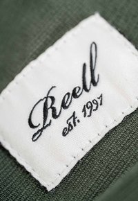 Etiqueta de tela blanca con texto bordado en negro "Reell est. 1997," unida a un material texturizado verde. Bordes cosidos visibles.