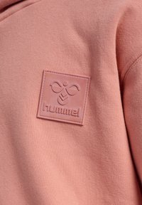 Pink stof med en syet firkantet lap med et præget bi-logo og ordet "hummel" på bagsiden af et tøjde.