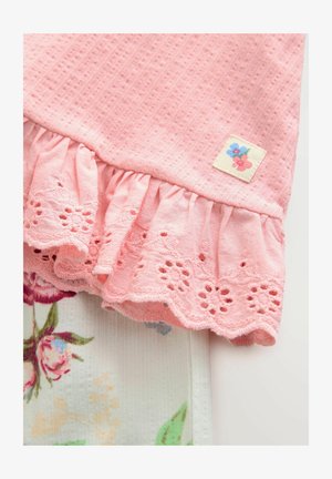 Roza pamučna odjeća s teksturiranim vrhom, volanastim rubom i čipkastim cvjetnim detaljima. Dijelom je složena preko tkanine s cvjetnim uzorkom.