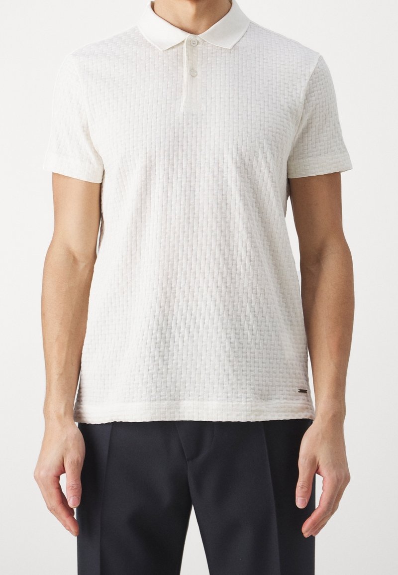 Polo shirt blanc à manches courtes avec texture côtelée, col et patte de boutonnage à trois boutons. Présente un logo discret au niveau de l'ourlet, associé à un pantalon foncé.