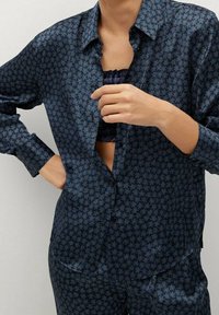 Persona che indossa una camicia a maniche lunghe con motivo a pois blu scuro e pantaloni abbinati, mostrando un top bandeau nero sottostante.