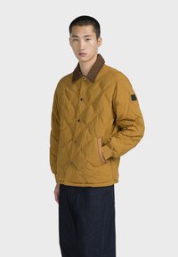 Giacca trapuntata gialla senape con colletto in velluto a coste marrone e accenti beige a contrasto. Presenta bottoni a pressione e tasche laterali.