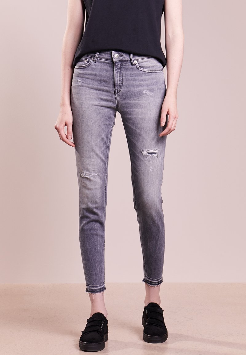 Jeans ajustados gris deslavado con detalles desgastados, cintura media y dobladillo sin acabar. Combinados con zapatos negros.