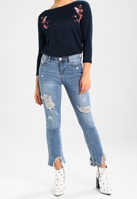 Vrouw in een marineblauwe top met bloemenborduursel, lichtblauwe gescheurde spijkerbroek met rafelige zoom en witte studded enkellaarsjes met hak.