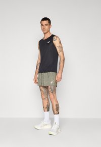 Svart ermeløs treningstopp, grønne mønstrede shorts, hvite sneakers med neonlysdetaljer, modell som viser ben-tatoveringer, står mot en nøytral bakgrunn.