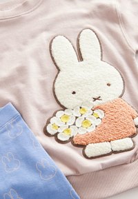 Ljust rosa sweatshirt med en texturerad vit kanindesign som håller blommor; blå byxor med upprepade ljusrosa kaninkonturer.