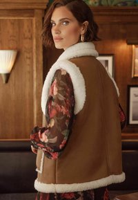 Gilet in suede marrone con colletto in pelliccia sintetica bianca, abbinato a un vestito lungo a maniche lunghe in chiffon floreale. Toni caldi e morbide trame visibili.