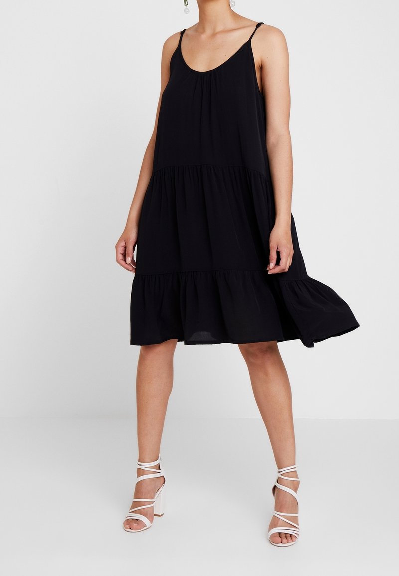 Femme portant une robe noire sans manches à étages et des sandales blanches à talons hauts à fines brides, debout devant un fond clair uni.