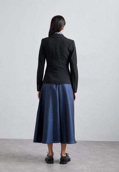 Svart figursydd kavaj i kombination med en flödande marinblå satinkjol, som har en lätt lyster och en slät textur. Svarta skor fulländar outfiten.