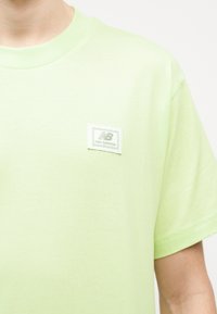 Άτομο που φοράει ανοιχτοπράσινο T-shirt με μικρό λευκό λογότυπο New Balance στο αριστερό στήθος, κοντά μανίκια και στρογγυλή λαιμόκοψη.