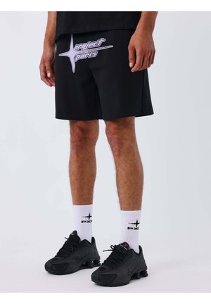 Hombre con pantalones cortos negros "Project Paris", calcetas blancas con logotipos negros y zapatillas de correr Nike negras, de pie sobre un suelo blanco.