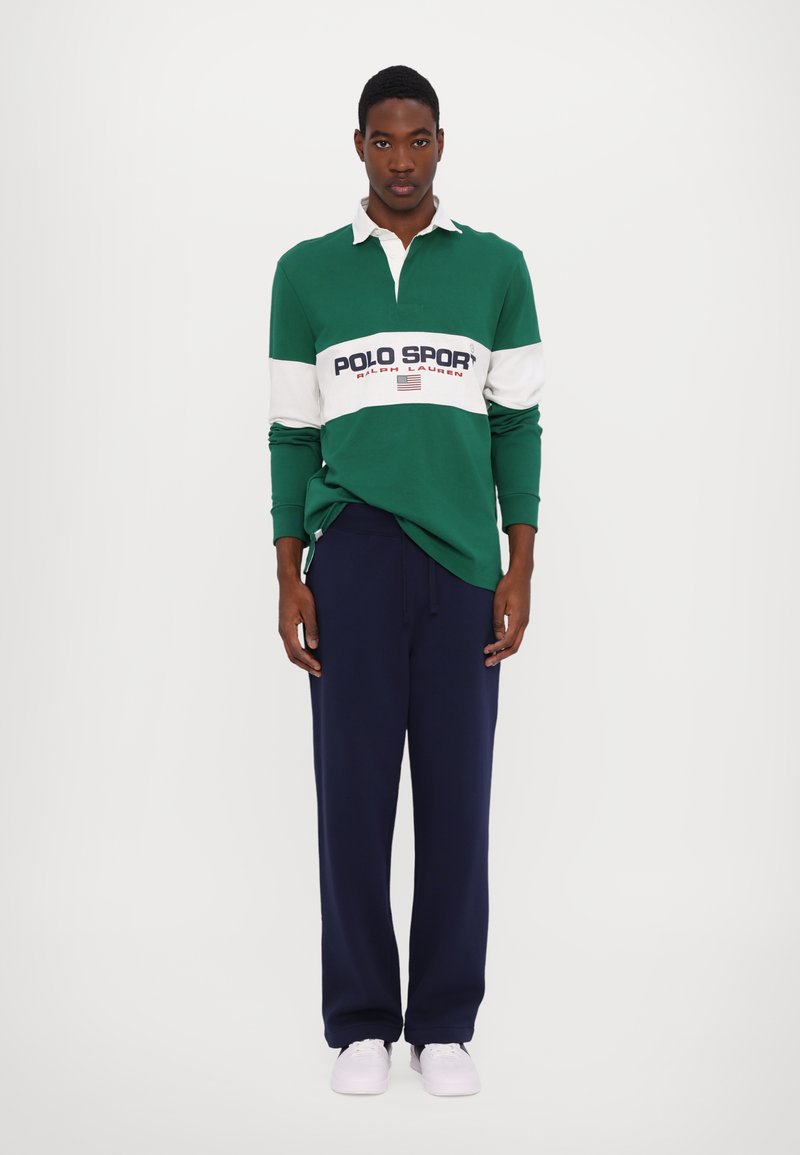 Grön långärmad polo med vit rand och texten "POLO SPORT", i kombination med marinblå träningsoverallsbyxor och vita sneakers. Slätt bomullstyg.
