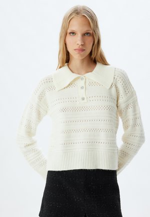 Pull en tricot blanc avec un ourlet côtelé, doté d'un grand col et de boutons décoratifs. Le motif texturé présente des rayures horizontales.