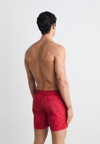 Shorts de bain rouges avec une taille élastique et une poche arrière, présentant une texture lisse et un design ajusté, vus de l'arrière.