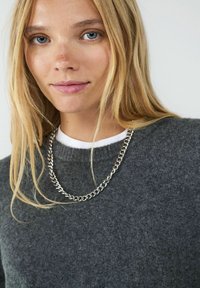 Grauer Wollpullover mit Rundhalsausschnitt, über einem weißen Shirt getragen, mit einer silbernen Halskette. Neutraler Hintergrund.