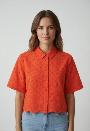 Femme aux cheveux bruns mi-longs portant une chemise orange à boutons ajourés et un jean bleu clair, debout devant un fond gris uni.