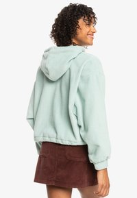 Sudadera de forro polar verde menta clara con capucha ajustable y hombros caídos, combinada con una falda de pana marrón con detalles de bolsillos.