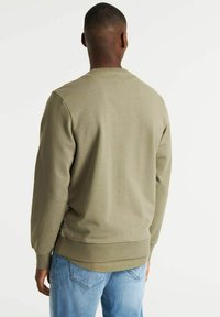 Felpa verde oliva con scollatura a girocollo, polsini e orlo a costine, dettagli di cucitura visibili, indossata con jeans azzurri chiari.