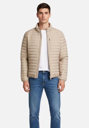 Jonge man die een beige gewatteerd jack draagt over een wit shirt en een blauwe spijkerbroek, staand tegen een effen achtergrond.