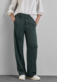 Osoba ubrana w luźne czarne spodnie z przednimi przeszyciami, białą koszulę na guziki z podwiniętymi rękawami oraz białe trampki, stojąca z rękami w kieszeniach.