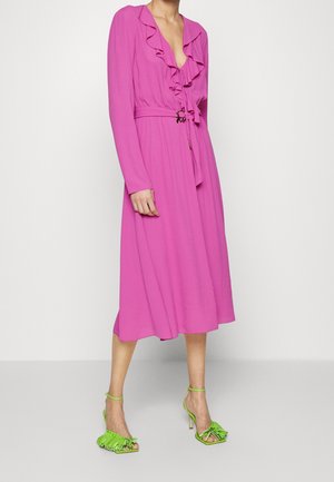 Woman wearing a long-sleeve, bright pink midi dress with ruffled neckline and tie belt, paired with neon green high-heeled sandals with fringe details.