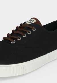 Baskets en toile noire avec des lacets marron, semelle en caoutchouc blanche et patch logo marron sur la languette, vues de côté et de face.