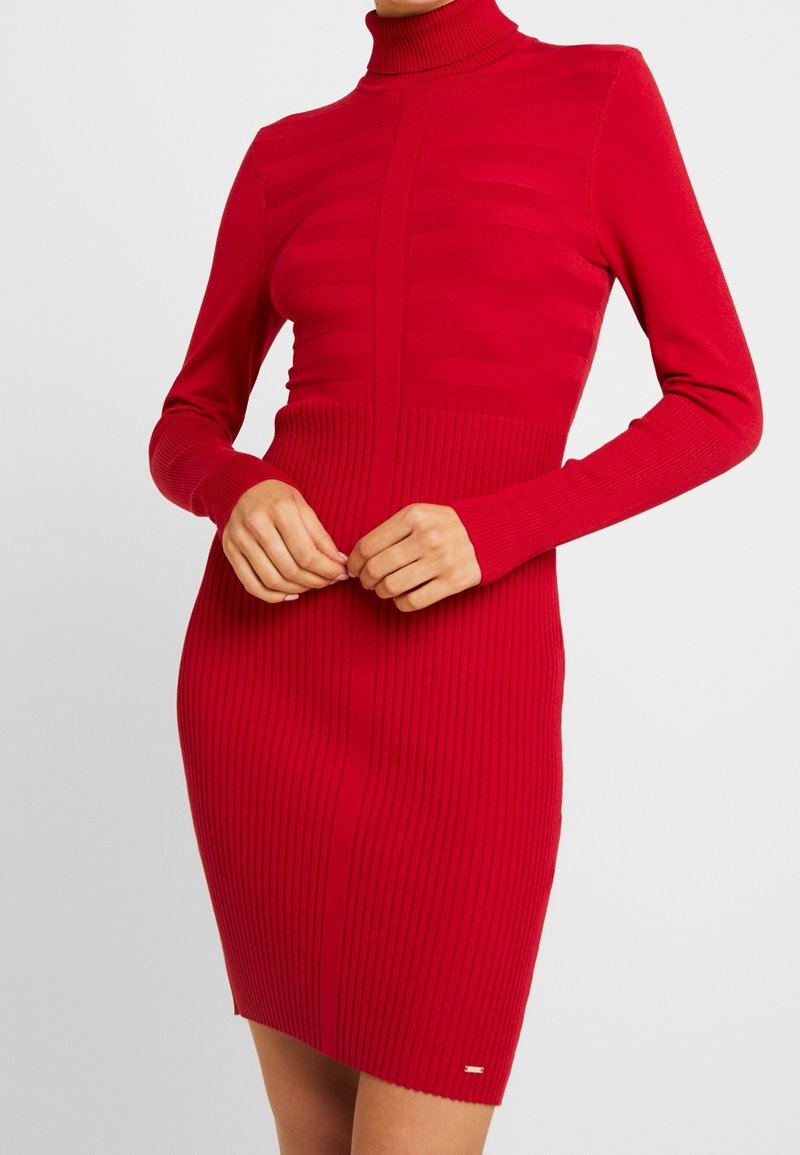 Femme portant une robe ajustée en maille côtelée rouge avec manches longues et col roulé, mains doucement jointes à la taille.