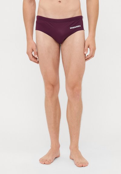 Man staat blootsvoets in maroon DSQUARED2 zwemboxers tegen een witte achtergrond, zichtbaar van taille tot voeten.