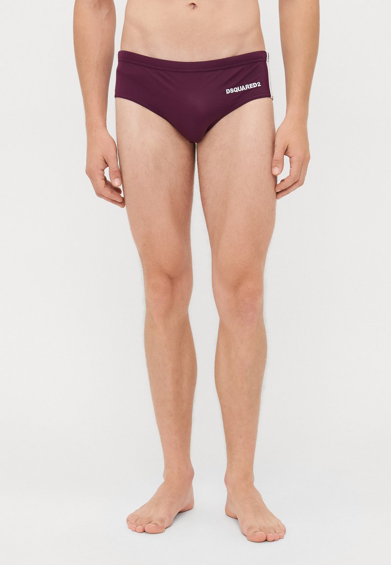 Mann steht barfuß in maroonfarbigen DSQUARED2 Badebriefs mit weißem Hintergrund, sichtbar von der Taille bis zu den Füßen.