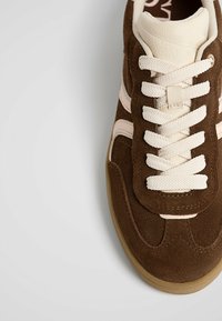 Brown suede sneaker with cream laces and side stripes, featuring a rubber sole and smooth leather accents. Textured surface visible.