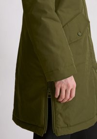 Grön vadderad jacka med dragkedja i sidan, tryckknapplåsning och en strukturerad ficka. Materialet verkar slätt med en något glansig yta.