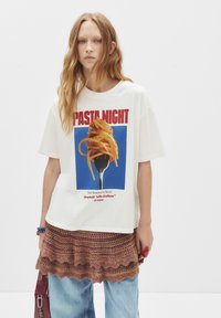 Mladá žena s dlouhými červenými vlasy, oblečená v bílém tričku "Pasta Night" s motivem špaget na vidlici, s pletenou sukní navrch a modrými džíny.