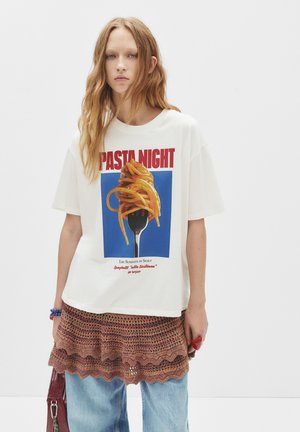 Jeune femme aux longs cheveux roux portant un T-shirt blanc "Pasta Night" avec des spaghetti sur une fourchette, une jupe en crochet par-dessus et un jeans bleu.