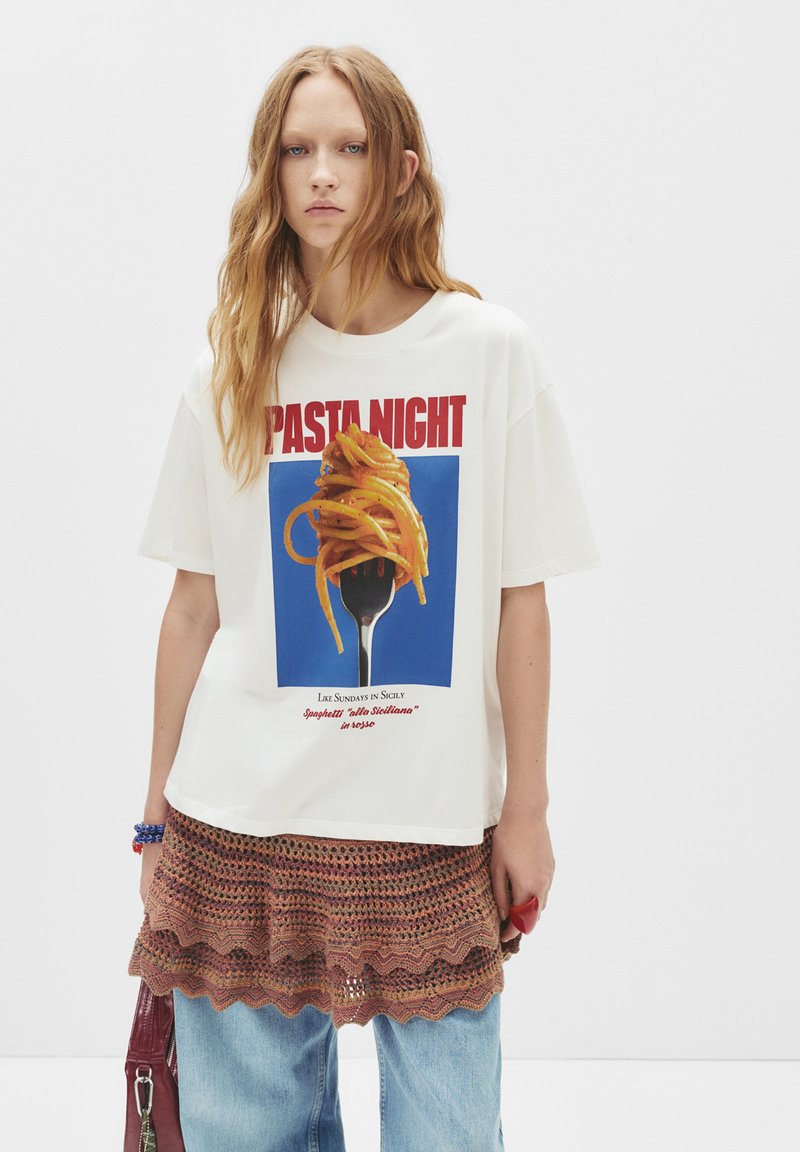 Mladá žena s dlouhými červenými vlasy, oblečená v bílém tričku "Pasta Night" s motivem špaget na vidlici, s pletenou sukní navrch a modrými džíny.